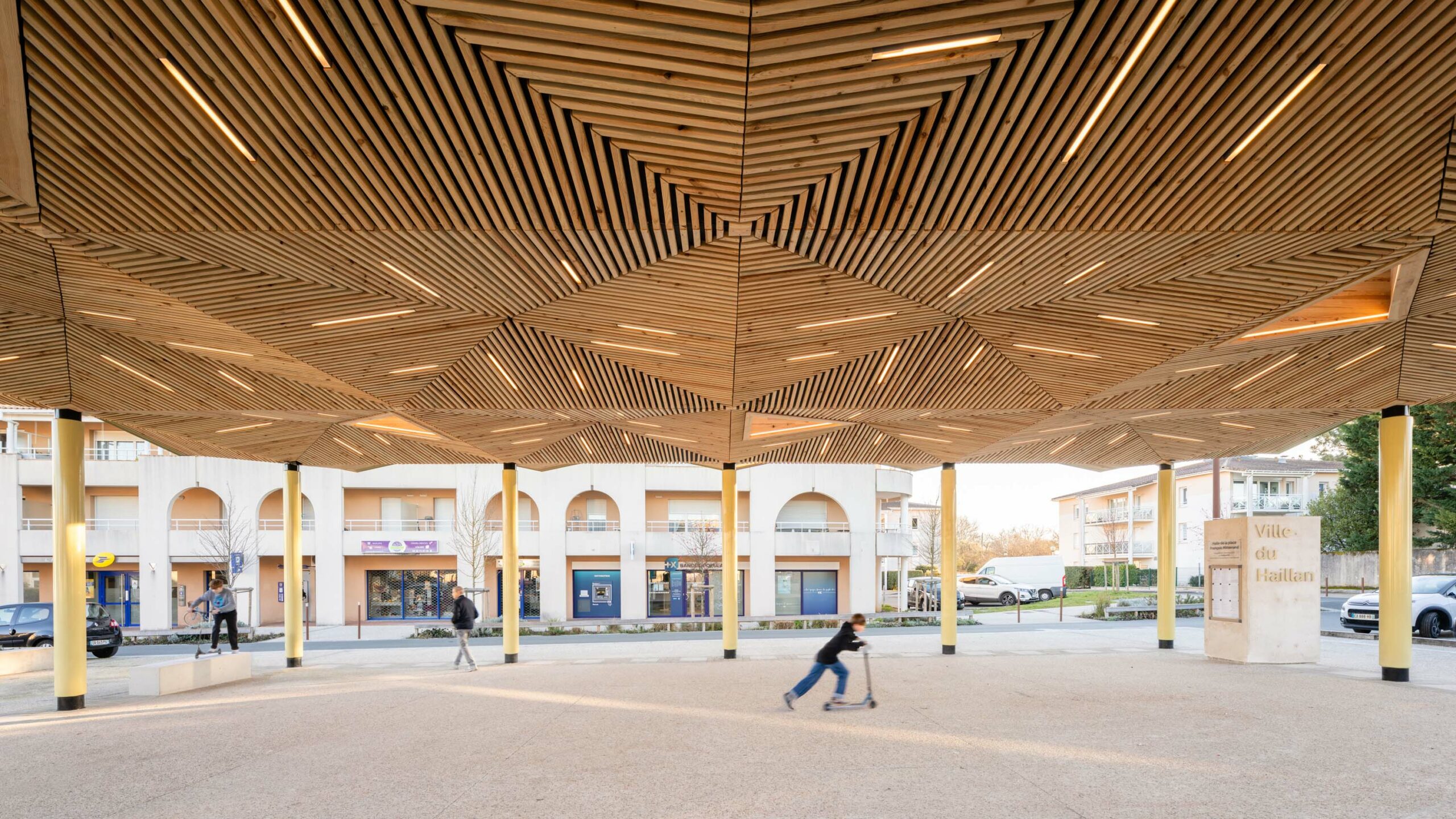 Vue depuis le dessous de la halle vers les commerces révélant le jeu de facettes de la sous face en bois de la halle / Florence VESVAL